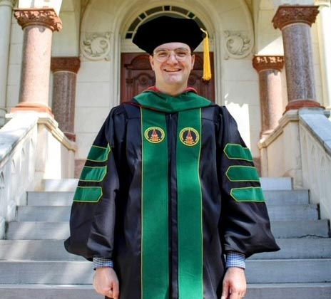 Jack Kennady in a graduation robe standing on steps in front of the school